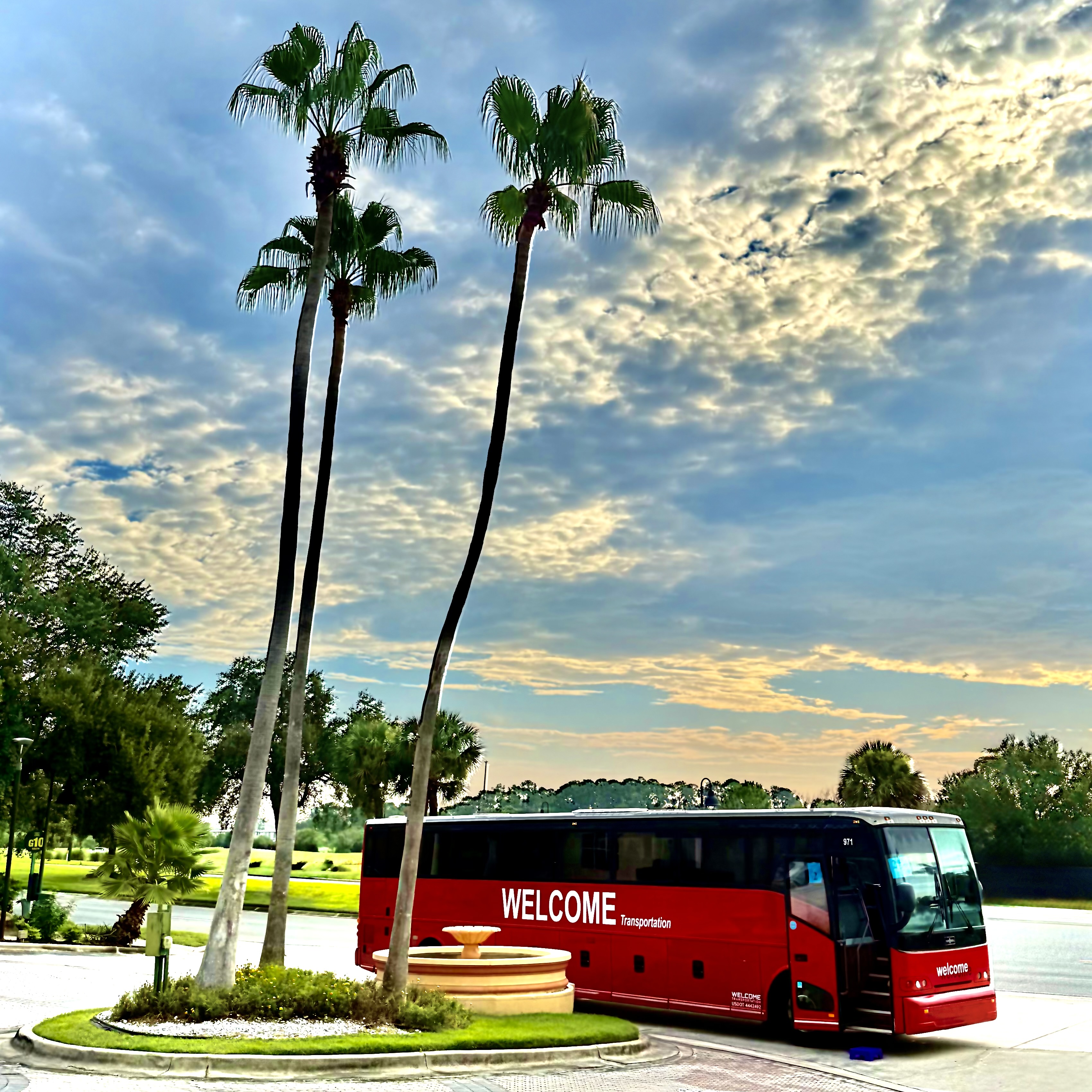 Welcome Transportation bus parked beside palm trees and a fountain at sunset
